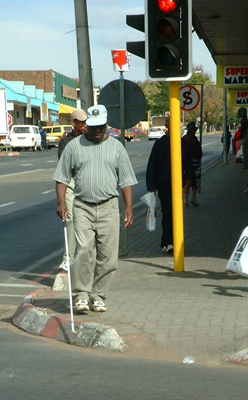 Photo shows a man wearing grey slacks and a soft-grey-and-green-striped shirt and baseball cap walking along a sidewalk and approaching a curb at a corner with a traffic signal.  He is reaching a long white cane with a bundu basher tip over the curb.